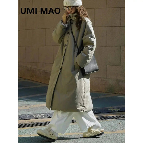 Woman walking outdoors in an army green long padded coat with a stand collar, styled with white trousers, a knit hat and a black shoulder bag