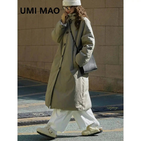 Woman walking outdoors in an army green long padded coat with a stand collar, styled with white trousers, a knit hat and a black shoulder bag