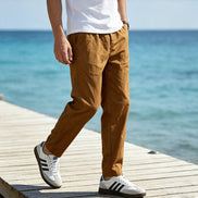 A person walking on a wooden dock by the ocean, wearing brown linen pants, a white t-shirt, and white sneakers with black stripes.