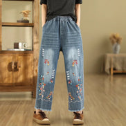 A person wearing loose, wide-leg blue denim jeans with colorful floral embroidery near the hem, paired with a dark gray top and brown shoes,