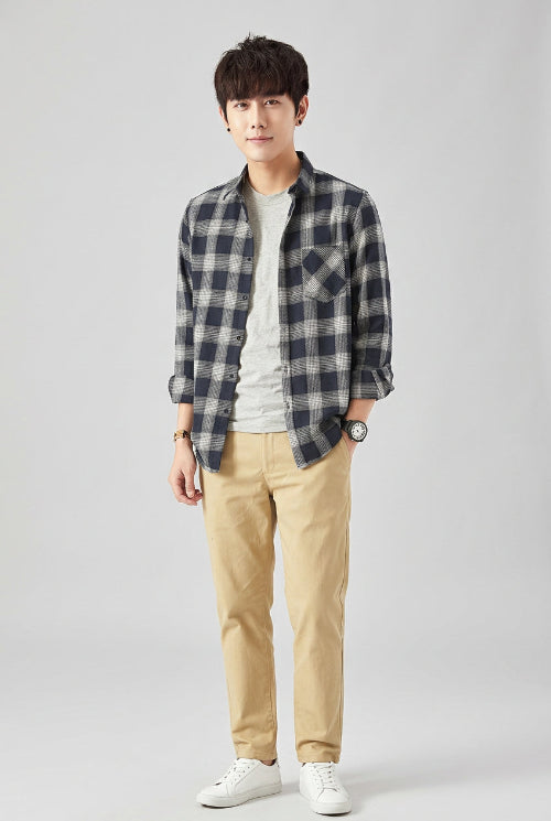 A male model wearing a dark navy and gray plaid button-down shirt over a light gray t-shirt and beige pants, 