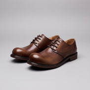 Pair of brown leather shoes on a wooden surface with vintage tools and a suitcase in the background.