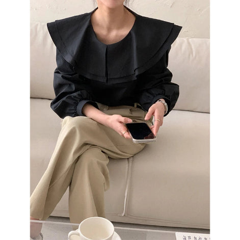 A woman seated on a light beige sofa wearing a black long-sleeve blouse with a dramatic oversized ruffled collar and beige wide-leg trousers, holding a smartphone.