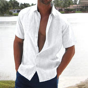 Man wearing a white short-sleeve shirt with open collar by a body of water.