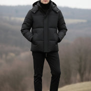 A man wearing a black hooded puffer jacket over a black shirt, shown from the neck down against a dark gray studio background. Three feature callout graphics appear on the left side, reading 85% DOWN, THICK WARM, and FLUFFY SOFT.