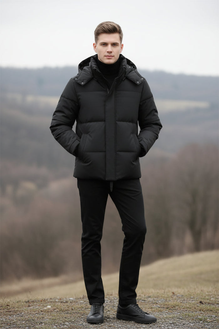A man wearing a black hooded puffer jacket over a black shirt, shown from the neck down against a dark gray studio background. Three feature callout graphics appear on the left side, reading 85% DOWN, THICK WARM, and FLUFFY SOFT.