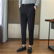 A person wearing slim black trousers with a button waist, paired with a gray long-sleeve shirt and black loafers, standing in an indoor room with wood flooring and a slatted wooden screen.