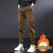 A person wearing brown corduroy jogger pants with a black top and white sneakers, standing against a dark studio background. 