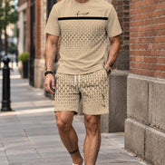 A man wearing a beige short-sleeve t-shirt and matching patterned shorts, leaning against a stone