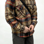 A person wearing a long-sleeve plaid button-up shirt in beige, brown, red, and navy tones, paired with dark pants and a black crossbody bag, against a plain white background.
