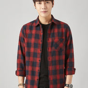 A man wearing a red and black plaid button-up shirt over a black t-shirt and black jeans, posing against a light gray background.