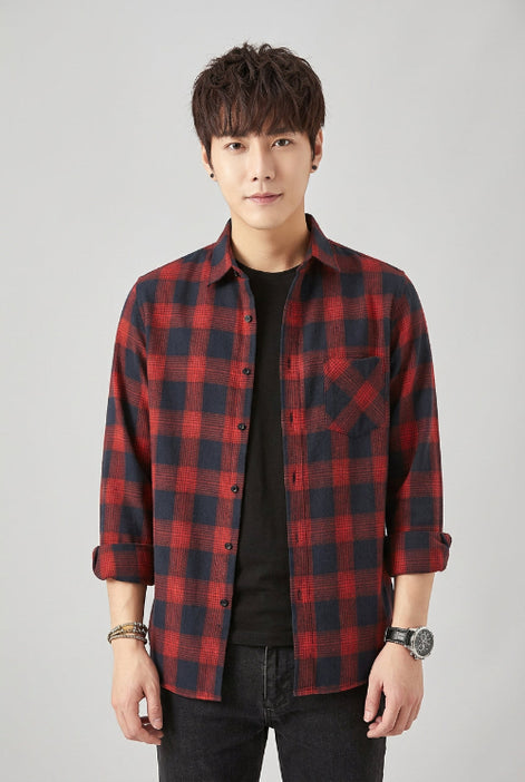 A man wearing a red and black plaid button-up shirt over a black t-shirt and black jeans, posing against a light gray background.