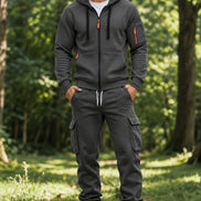 A man wearing a dark gray zip-up hoodie and matching jogger pants with cargo pockets, standing in a sunlit forest clearing.