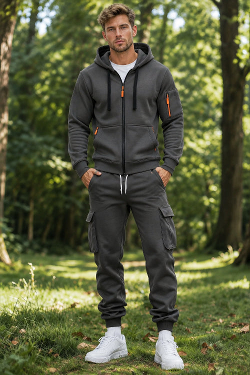 A man wearing a dark gray zip-up hoodie and matching jogger pants with cargo pockets, standing in a sunlit forest clearing.
