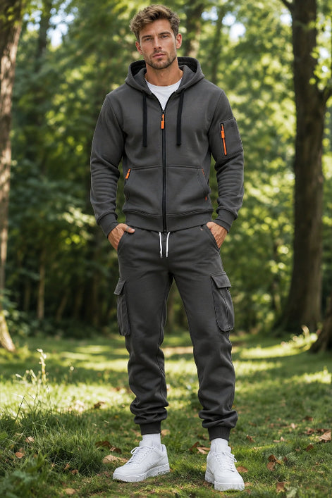 A man wearing a dark gray zip-up hoodie and matching jogger pants with cargo pockets, standing in a sunlit forest clearing.