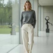 A woman wearing a gray long-sleeve knit sweater and black pants, standing indoors in front of white curtains.
