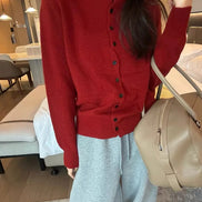 A person wearing a red mock-neck button-up knit sweater and light gray sweatpants, carrying a beige shoulder bag in an indoor room setting.
