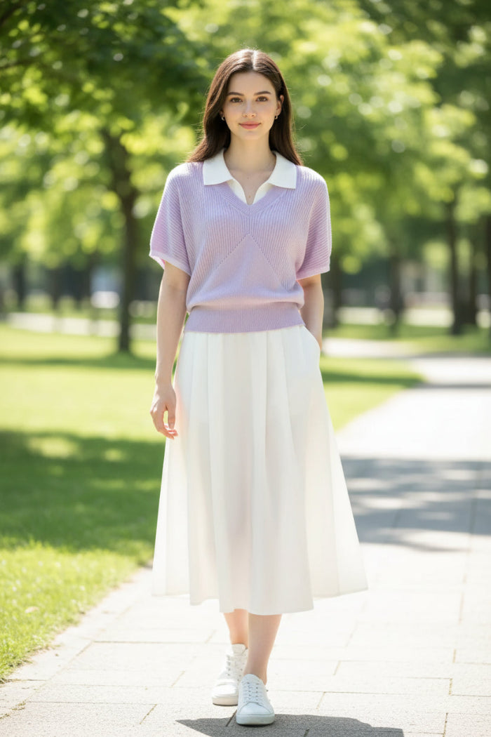 A woman wearing a light purple short-sleeve ribbed knit polo sweater over a white collared shirt, paired with a black polka-dot skirt and carrying a black shoulder bag.