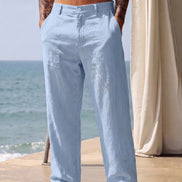 A man wearing light blue wide-leg linen pants, standing barefoot on a patio with the ocean and a cream-colored curtain in the background.
