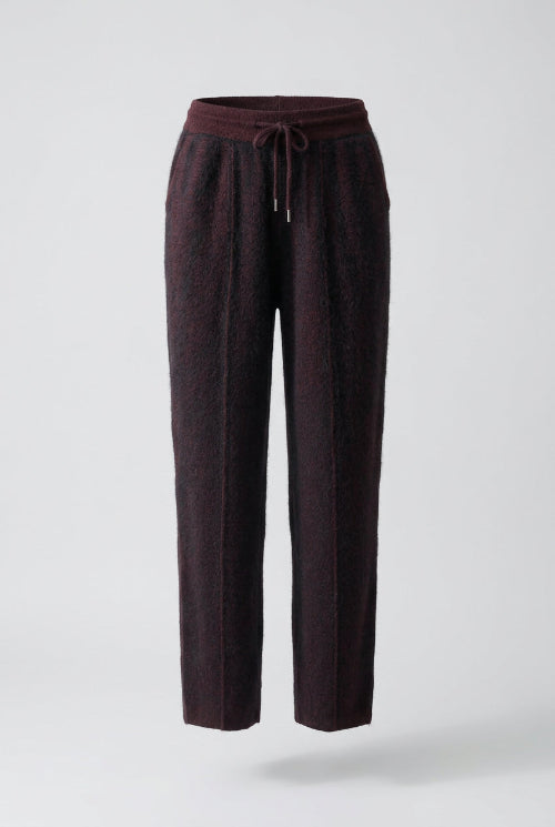Dark plum-colored knit pants with a drawstring waist, hanging on a wooden hanger inside an open wooden wardrobe or closet.