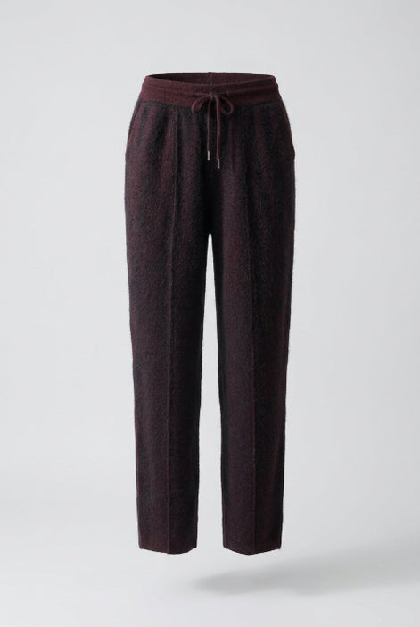 Dark plum-colored knit pants with a drawstring waist, hanging on a wooden hanger inside an open wooden wardrobe or closet.