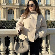 Woman wearing a beige double-breasted short coat while holding a handbag beside a stone railing