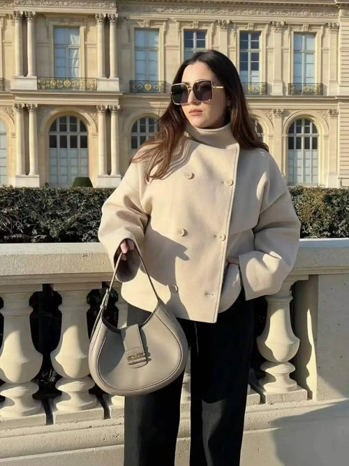 Woman wearing a beige double-breasted short coat while holding a handbag beside a stone railing