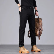A man wearing slim-fit black jeans, a black long-sleeve shirt, and tan lace-up boots, holding a brown leather backpack against a plain gray studio background.