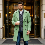 A man wearing a light green double-breasted overcoat over a gray shirt and blue jeans, holding a coffee cup in front of a hotel revolving door.