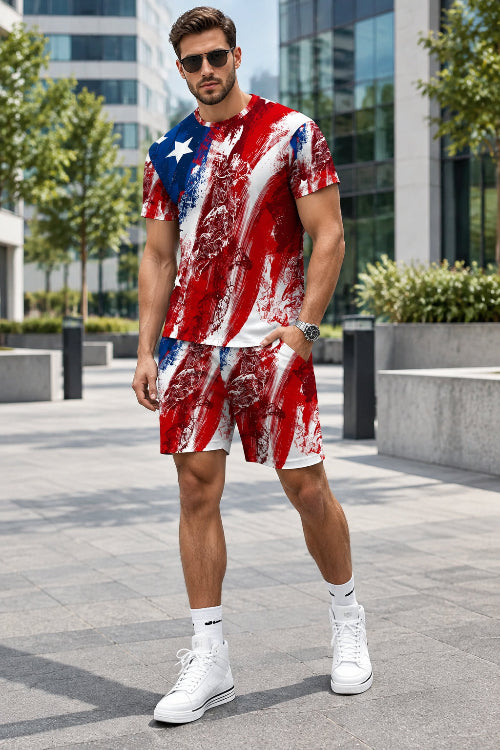 A man modeling a matching red, white, and blue distressed American flag t-shirt and shorts set, paired with white socks and sneakers, standing against a gray paneled wall.
