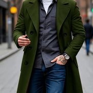 A man wearing a double-breasted olive green overcoat over a dark gray button-up shirt, blue jeans, and a white t-shirt, walking on a city street.