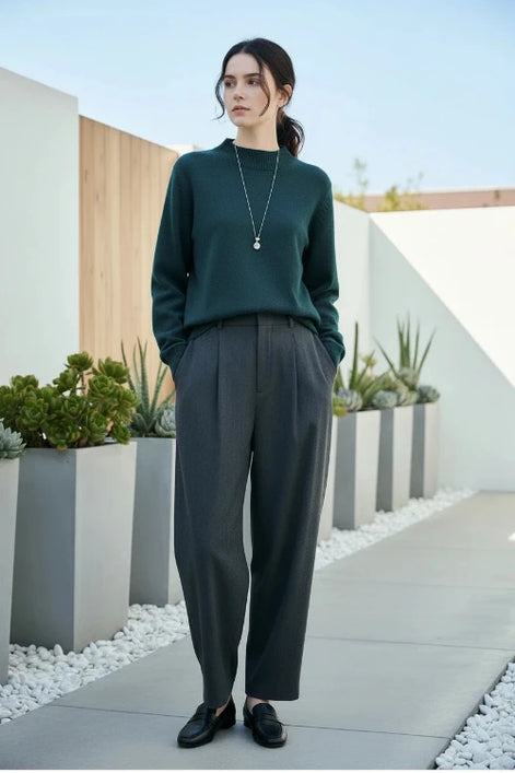 A woman wearing a dark gray ribbed knit mock neck sweater and black pants, sitting on a brown leather chair and wearing a silver pendant necklace.