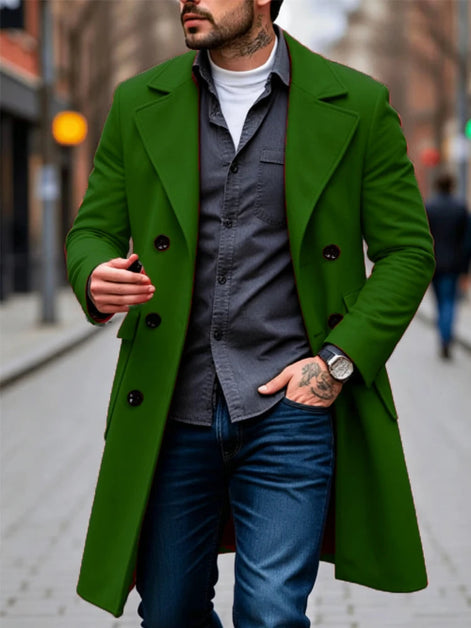 A man wearing a vibrant green double-breasted overcoat over a gray button-down shirt, white t-shirt, and blue jeans, walking on a blurred city street.