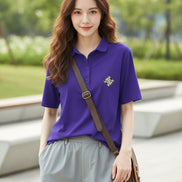 A woman wearing a short-sleeve purple polo shirt with a small white embroidered geometric logo on the left chest, paired with light gray bottoms and carrying a brown shoulder bag.