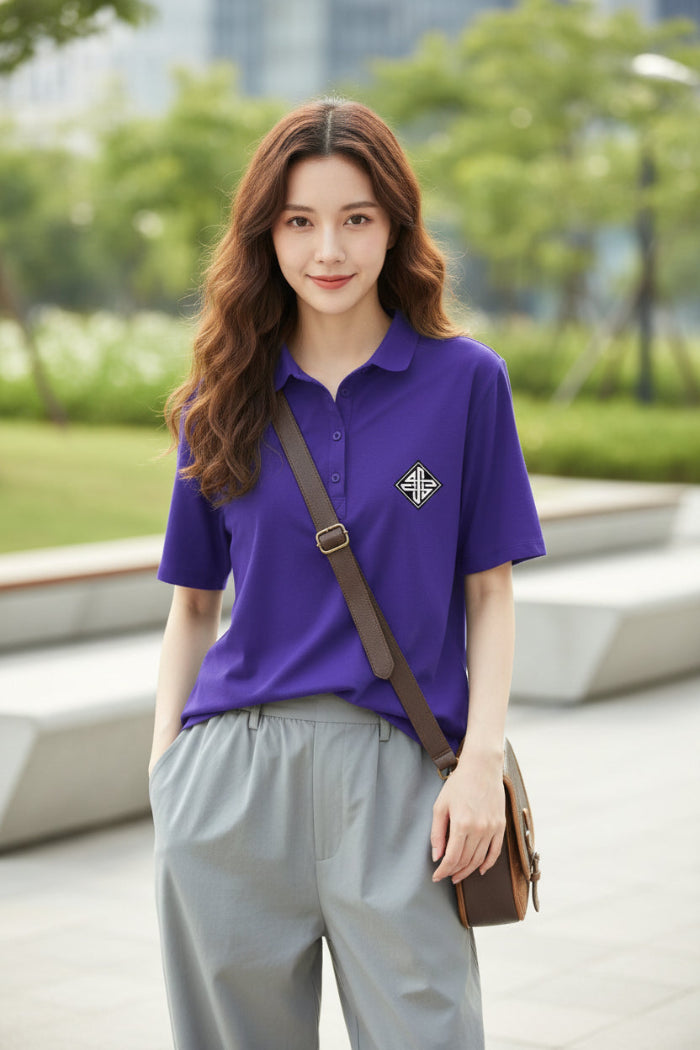 A woman wearing a short-sleeve purple polo shirt with a small white embroidered geometric logo on the left chest, paired with light gray bottoms and carrying a brown shoulder bag.