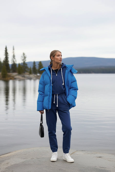 Person wearing a blue puffer jacket and tracksuit standing against a building.