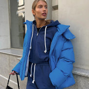 Person wearing a blue puffer jacket and tracksuit standing against a building.