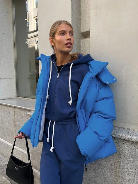 Person wearing a blue puffer jacket and tracksuit standing against a building.