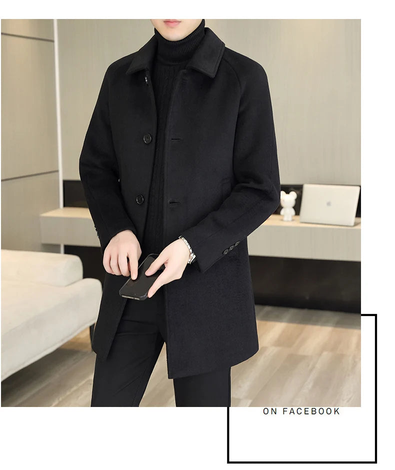 A man wearing a black mid-length coat over a black turtleneck sweater and black pants, standing in a modern indoor room with a bed and desk visible in the background.