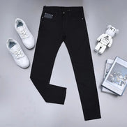 Black slim-fit jeans laid flat on a light gray surface, styled with white sneakers, a white bear figure, and a stack of magazines.