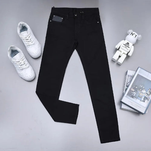 Black slim-fit jeans laid flat on a light gray surface, styled with white sneakers, a white bear figure, and a stack of magazines.