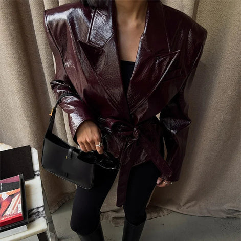 Angled close-up of a woman wearing a glossy dark brown jacket with wide lapels and a tied waist belt, holding a black handbag beside curtains⠋
