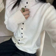 A person wearing a white ribbed turtleneck cardigan sweater with large dark buttons, paired with dark pants, sitting indoors.