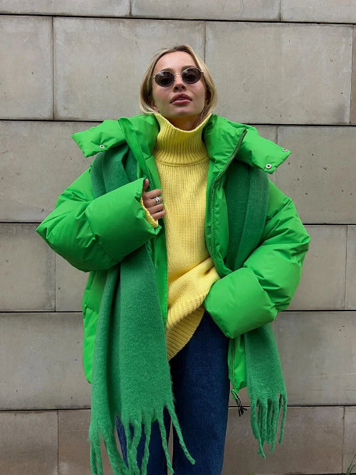 Person wearing a bright green coat with a yellow sweater and blue jeans against a stone wall.
