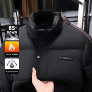 A black puffer jacket with a high collar and snap-button front, shown hanging on a hanger with a hand holding the fabric to demonstrate its soft, warm texture. Three promotional graphic overlays on the left highlight features: '85% DOWN', 'THICK WARM', and 'FLUFFY SOFT'.