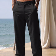 A man wearing black wide-leg trousers stands barefoot on a patio with the ocean and a cream curtain in the background.