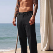 A man wearing black wide-leg trousers stands barefoot on a patio with the ocean and a cream curtain in the background.