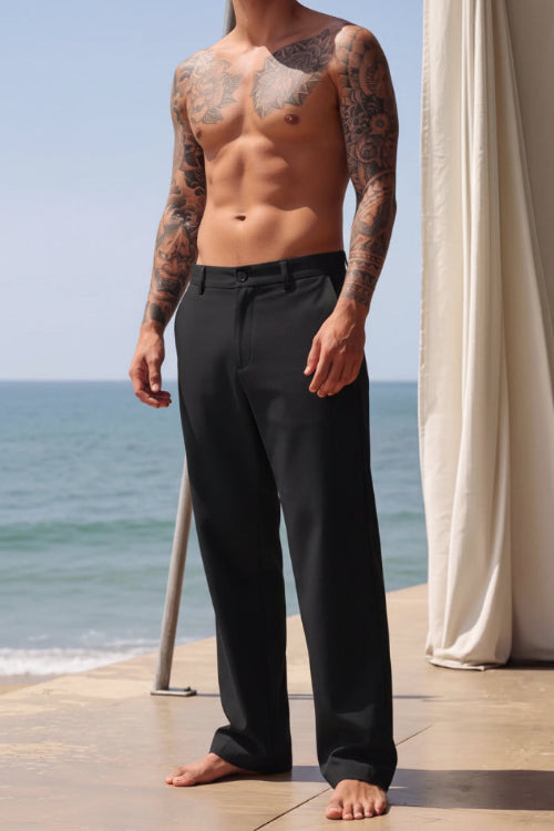 A man wearing black wide-leg trousers stands barefoot on a patio with the ocean and a cream curtain in the background.