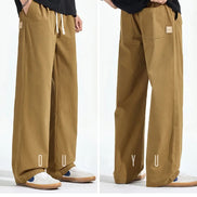 Front and back views of a model wearing khaki loose-fit straight-leg pants, showing a white drawstring, side pockets, and back patch pockets with a small sewn-on label.