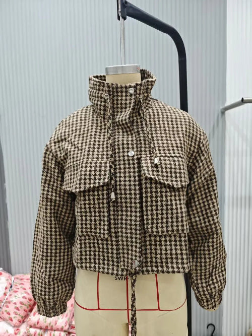 Brown and cream houndstooth utility jacket on a dress form, showing a raised collar, chest flap pockets, front snap buttons, elasticated cuffs and a drawcord hem